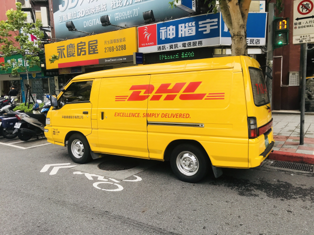 Ein gelber DHL-Lieferwagen steht auf der Straße neben einem Gebäude, umgeben von Bäumen, Schildern, Pfählen, Verkehrsschildern und einer Person auf dem Gehweg.