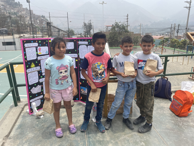 Gruppe von Kindern auf einem Basketballfeld mit Papiertüten in den Händen, mit Tüten und Gegenständen rechts und einer Tafel mit Text links, vor einem Hintergrund aus Zäunen, Masten, Straßlen, Bäumen, Gebäuden, Hügeln und Himmel.