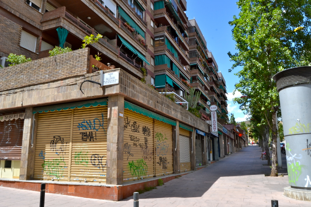 Eine Szene mit mehreren Gebäuden mit Fenstern, grünen Kleidungsstüccken, Geschäften, Schildern und umgebenden Bäumen und Pflanzen.