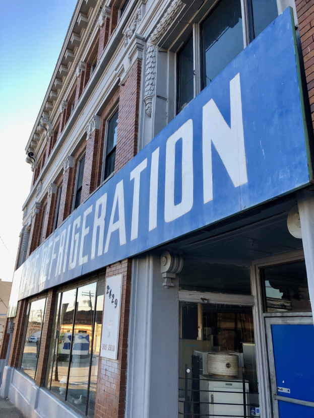 Ein modernes Gebäude mit Glaswänden und einem blauen Schild, das einen sichtbaren Kühlschrank enthält, vor einem Himmelhintergrund.