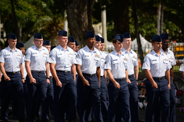 Männer in Uniformen gehen auf einer Straße mit einem Eisengeländer dahinter, Bäume und Pfähle im Hintergrund.