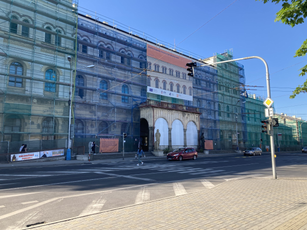 Großes Gebäude mit Gerüst bei Renovierung, umgeben von Fahrzeugen, Fußgängern, Ampeln, Schildern, Straßeninfrastruktur, Bäumen und einem klaren blauen Himmel.