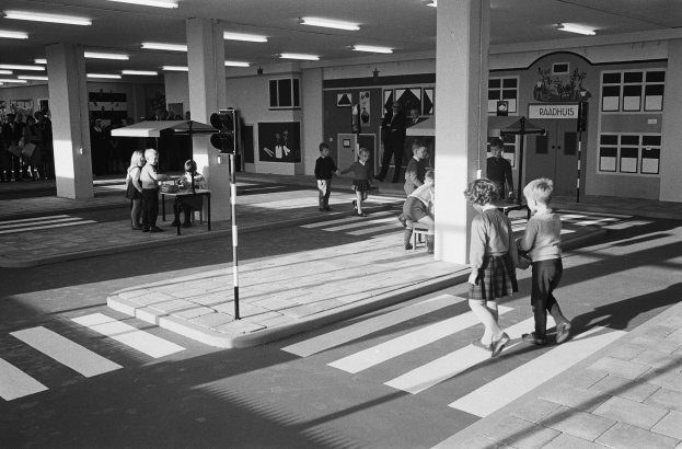 Schwarz-weiß-Foto von Kindern, die eine Straße überqueren, mit Passanten in der Nähe, einem Verkehrszeichenmast im Vordergrund und Hintergrundsäulen mit Deckenlampen.