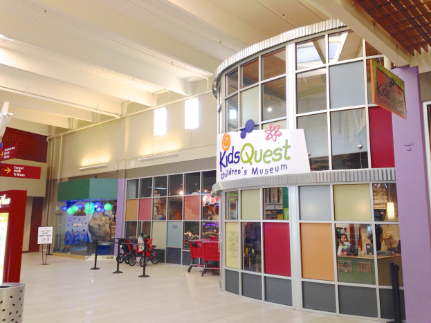 Innenansicht eines Einkaufszentrums mit einem "Kids Quest Children's Museum"-Schild, mit Trolleys, ribbon-adorned poles, informational boards, ceiling lights und verschiedenen verstreuten Gegenständen.