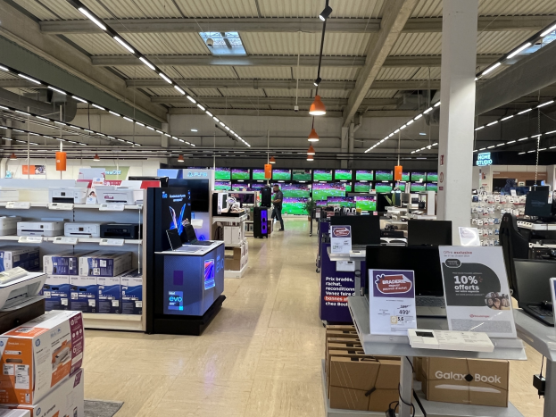 Ein großer Elektronikbereich in einem Walmart-Laden mit Regalen voller Computer, Drucker und Kartons, mit Menschen, die durch die Gänge schlendern, Deckenventilatoren, Lampen und Informationsschilder.