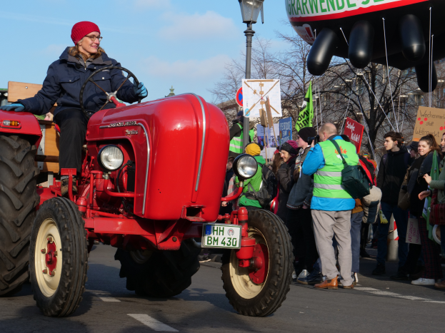 Eine Frau fährt einen roten Traktor auf einer Straße, umgeben von einer Menge mit Schildern und Transparenten, mit Laternen, Bäumen, Gebäuden und einem klaren blauen Himmel im Hintergrund.