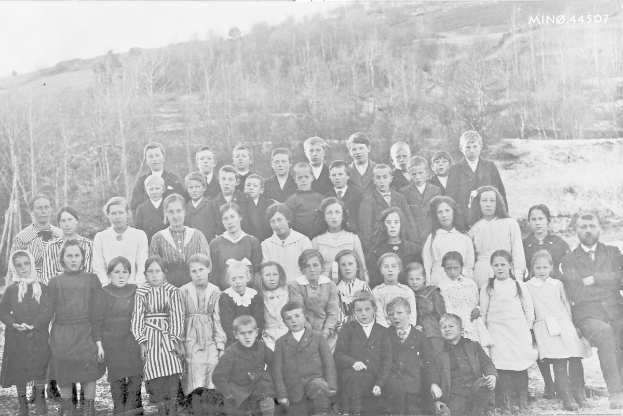 Ein Schwarz-Weiß-Foto einer Gruppe von Kindern, die vor Bäumen und einem Hügel posieren, mit dem Text "1930-1940 Schulkinder" unten.