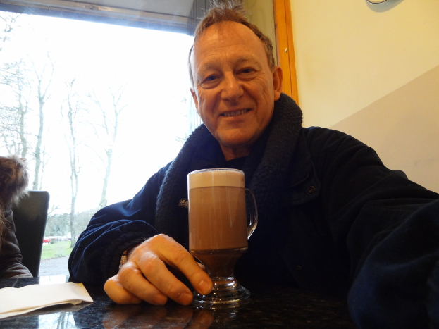 Ein Mann sitzt an einem Tisch mit einem Bierglas vor ihm, in einem Raum mit Bäumen und Gras draußen.