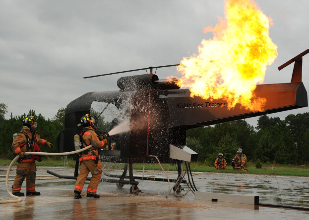 Feuerwehrleute mit Helmen spritzen Wasser auf einen brennenden Hubschrauber, während im Hintergrund Bäume und ein klarer Himmel zu sehen sind.