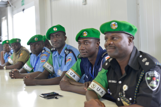 Eine Gruppe von Polizeibeamten sitzt an einem Tisch mit Mobiltelefonen und anderen Gegenständen in einem Raum mit einer Wand, Fenstern links und einer Decke mit Lampen.