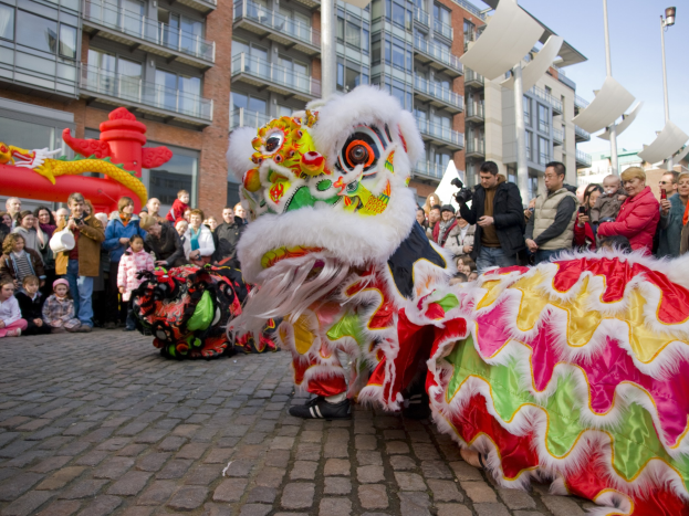 Ein lebendiges chinesisches Neujahrsfest in Amsterdam mit einem Löwen tanzen im Vordergrund und einer Menschenmenge drumherum, einige halten Kameras in den Händen, und im Hintergrund sind Gebäude, Laternenmasten und ein klarer blauer Himmel zu sehen.