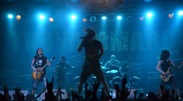 Eine Gruppe von Musikern, die auf einer hell erleuchteten Bühne mit Gitarren, Mikrofon und Trommeln spielen, während das Publikum im Vordergrund die Hände hebt.