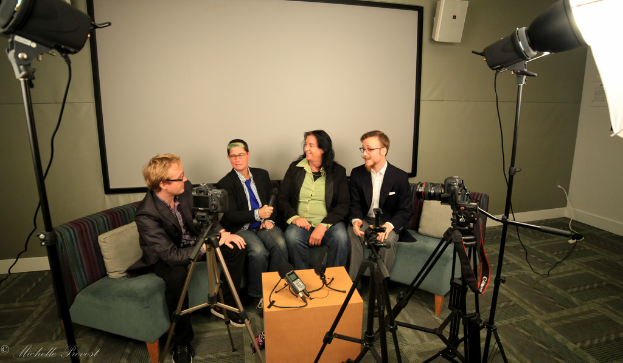 Eine Gruppe von drei Personen sitzt auf einer Couch und gibt ein Interview, wobei eine Person links in einem schwarzen Mantel und Hose, eine Person in der Mitte in einem Mantel und blauer Krawatte mit einem Mikrofon und eine Person rechts mit einem strahlenden Gesichtsausdruck zu sehen ist, sowie ein Projektionsschirm dahinter, ein Mikrofonrekorder auf einem Tisch in der Mitte und Kameras mit großen Stativen auf beiden Seiten.