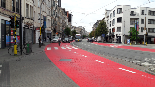 Eine Stra√e in der Stadt mit einem roten Fahrradweg, fahrende Fahrzeuge, Passanten auf dem Gehweg, Gebäude mit Fenstern auf beiden Seiten und Bäume mit einem klaren blauen Himmel im Hintergrund.