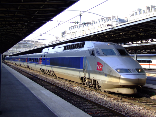 Hochgeschwindigkeitszug fährt auf Gleisen neben einem Bahnsteig mit Gebäuden und einem klaren blauen Himmel im Hintergrund.