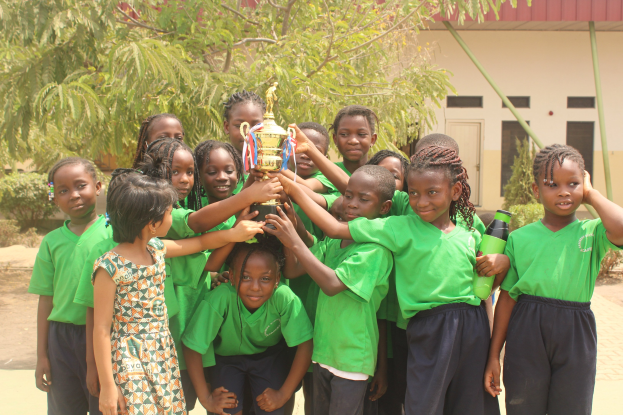 Gruppe von Kindern mit einem Pokal in einer Schulumgebung mit Bäumen und einem Gebäude im Hintergrund.