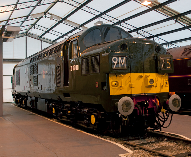 Ein Zug fährt auf den Schienen neben einem Bahnsteig mit einem Mast auf dem Dach, an dem Metallstangen mit Lampen befestigt sind.