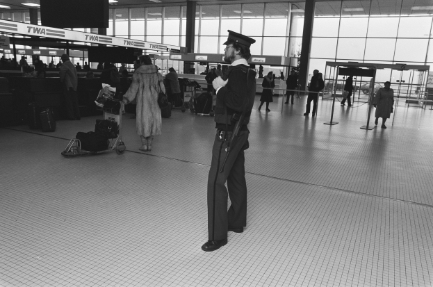 Polizeibeamter in Uniform mit Pistole in einem Flughafen-Terminal umgeben von Menschen, Gepäckwagen und Flughafen-Infrastruktur.
