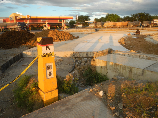 Eine Baustelle mit einem gelben Warnschild am Straßenrand, umgeben von Gras, Steinen, Pflanzen und einem gelben Band an einem Pfahl, mit Fahrzeugen, einem Gebäude, Bäumen, Pfählen, Drähten und einem bewölkten Himmel im Hintergrund.