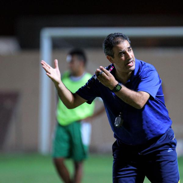 Ein Mann in einem blauen T-Shirt und Hose steht auf einem Fußballfeld und hält ein Mikrofon in der Hand, während er zu seinen Spielern zeigt. Im Hintergrund sind eine Person in einem grünen Kleid, ein Torpfosten und eine Wand zu sehen.