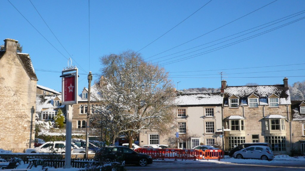 Fahrzeuge auf einer schneebedeckten Straße mit schneebeladenen Häusern, Bäumen, Strommasten und Leitungen im Hintergrund.