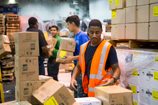 Eine Gruppe von Männern in orangenen Westen und Sicherheitswesten lädt Kisten in ein Lagerhaus, mit mehreren Kartonagen auf der rechten Seite und einer Kette auf der linken Seite und einer beleuchteten Wand im Hintergrund.