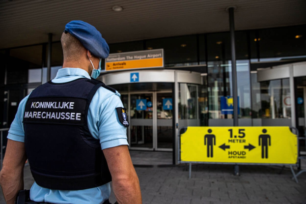 Ein Polizist in blauer Uniform und Mütze steht vor einem Glasgebäude, trägt eine Maske und befindet sich in der Nähe eines gelben Schildes mit Text.
