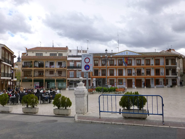 Ein belebter Stadtplatz mit Menschen auf Stühlen sitzend und stehend, umgeben von Topfpflanzen, Metallabsperrungen, Straßenlaternen mit Fahnen, einem Schild, Gebäuden mit Fenstern und einem bewölkten Himmel.
