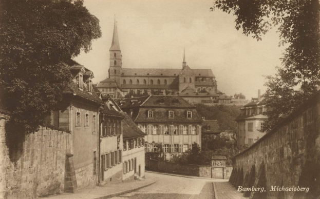 Schwarz-weiß-Foto einer Stadtstraße mit einer Kirche im Hintergrund, Mauern auf beiden Seiten, Bäume entlang der Straße und Himmel darüber; die Kirche befindet sich in Bamberg, Deutschland.