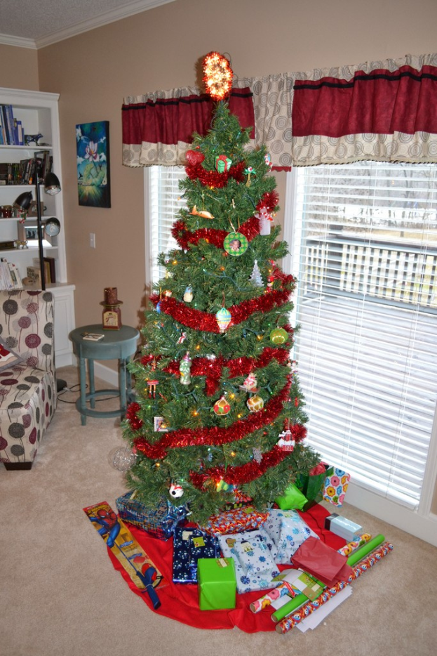 Ein Weihnachtsbaum steht neben einem Fenster mit Vorhängen, einem Bücherregal, einer Stehlampe, einem Sofa, einem Tisch, einem Bild an der Wand und Geschenken vor dem Baum.