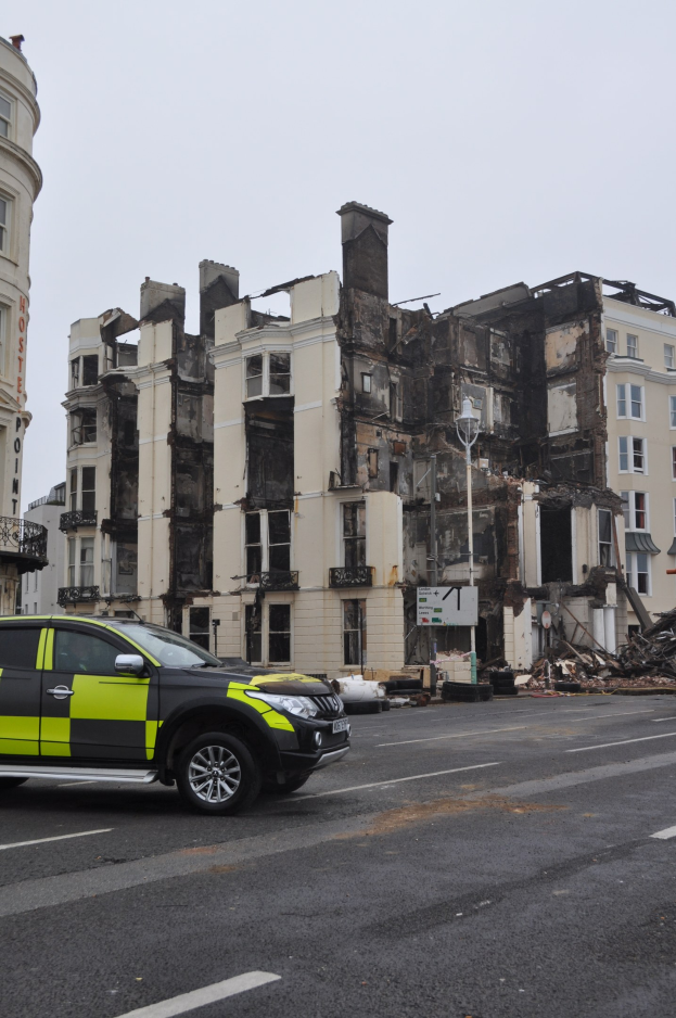 Ein vor einem schwer beschädigten Gebäude geparkter Polizeiwagen mit zersplitterten Fenstern und verstreuter Trümmer, unter einem sichtbaren Himmel mit benachbarten Gebäuden.