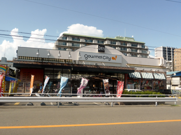 Eine belebte Stadtstraße mit Gebäuden, Straßeneinrichtungsgegenständen, Fahrzeugen, Grünflächen und einem Restaurant, das Speisen in seinem Fenster präsentiert.