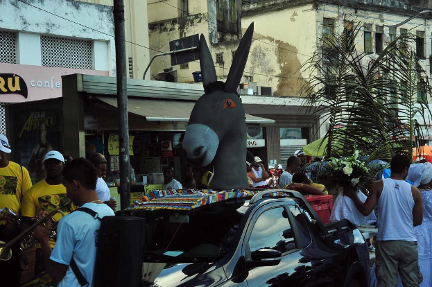 Eine Skulptur in der Mitte mit Menschen drumherum, Gebäuden und Drähten im Hintergrund, ein Blumenstrauß, ein Auto, ein Pfahl, eine Person, die auf der linken Seite ein Musikinstrument spielt, und ein Geschäft.
